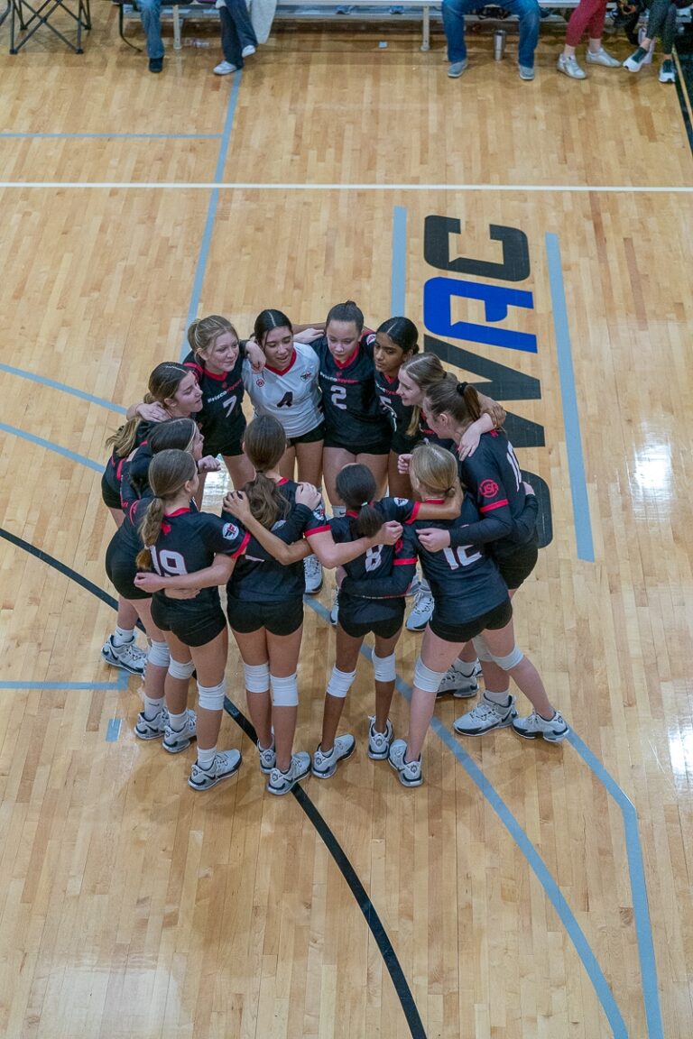 A women's volleyball team stands in a tight huddle on an indoor court, wearing matching black uniforms with red accents. The wooden floor has blue and black markings, setting the stage for the U12 Season of 2024.