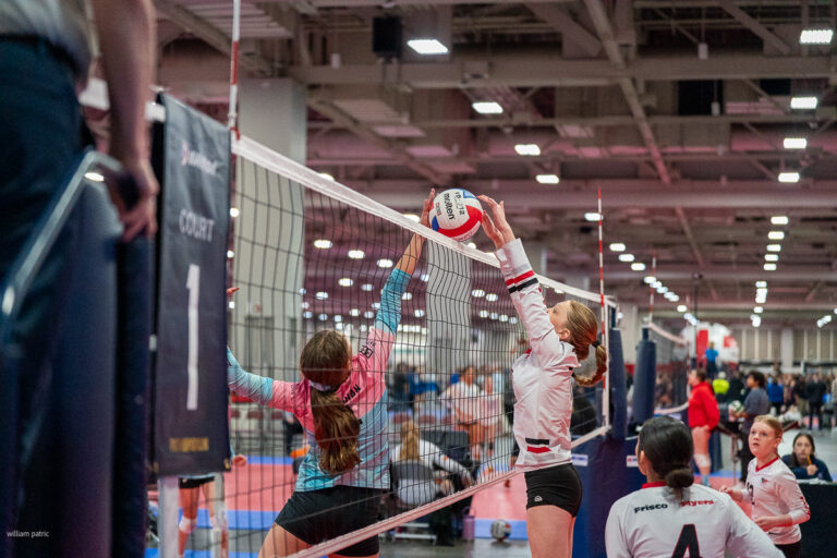 Two volleyball players at the net, one in a pink jersey attempting to block while the other in a white jersey hits the ball, with teammates and spectators in the background.