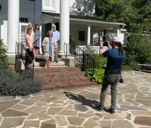 A photographer is taking a picture of five people standing on the steps of a white building with columns. The group, curated by charlie patric photography, poses elegantly on a stone patio with lush greenery in the background.