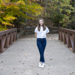 A person stands on a leaf-covered bridge with hands behind their back, wearing a white shirt and jeans, surrounded by trees with autumn foliage—a picturesque moment straight out of a Charlie Patric photography video.