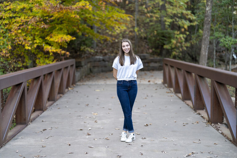 A person stands on a leaf-covered bridge with hands behind their back, wearing a white shirt and jeans, surrounded by trees with autumn foliage—a picturesque moment straight out of a Charlie Patric photography video.