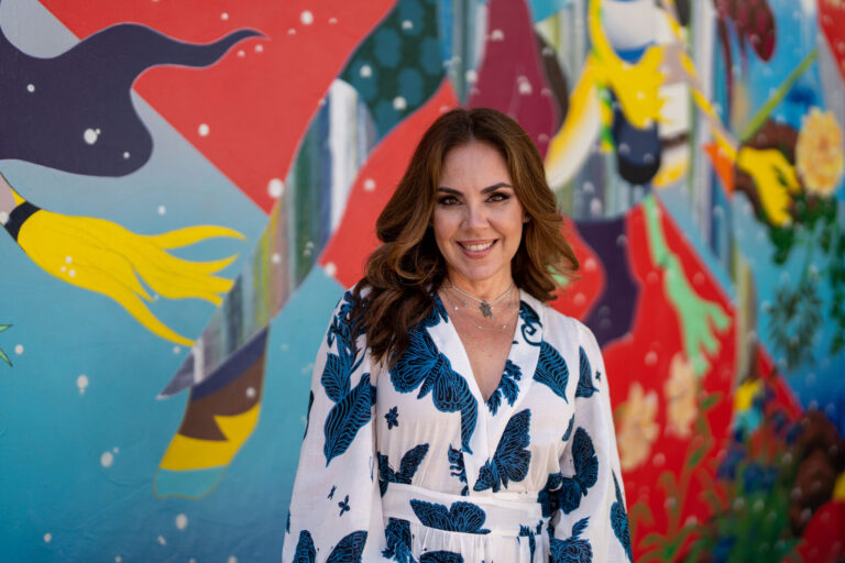 A woman in a white dress with blue patterns stands in front of a colorful mural, as Charlie Patric captures the moment with his keen eye for video and photography.