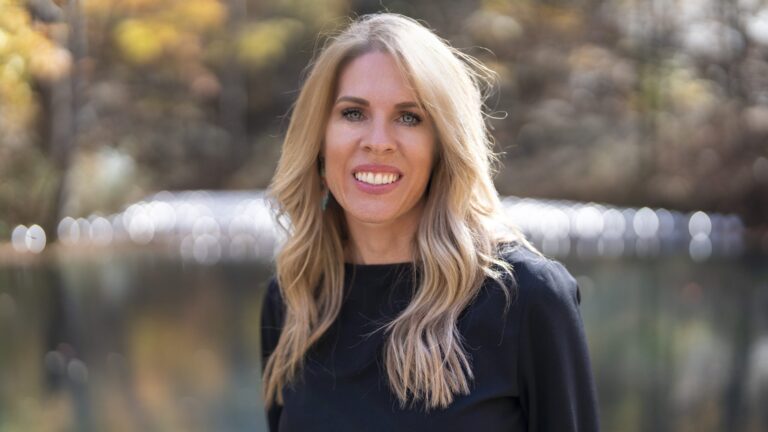 A woman with long blonde hair and a black top smiles at the camera, capturing a moment of joy while standing outdoors with a blurred natural background, perfect for any Charlie Patric video or photography project.