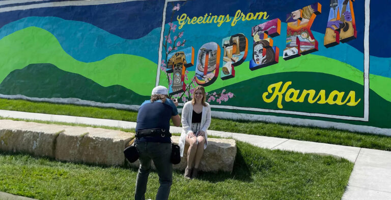 A woman poses for a photo in front of a colorful mural that reads "Greetings from Topeka, Kansas," while a photographer captures the moment. It’s clear that Charlie Patric’s keen eye for photography transforms it into an unforgettable image.