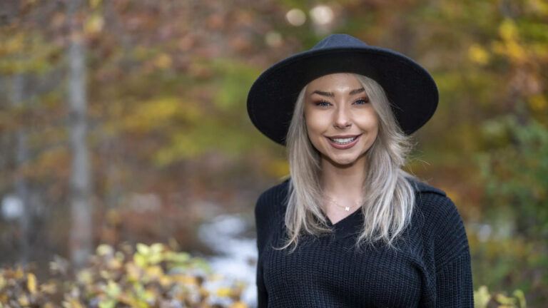 A woman with long blonde hair wearing a black hat and a black sweater stands outdoors, captured in stunning photography by Charlie Patric, against a blurred background of autumn foliage.