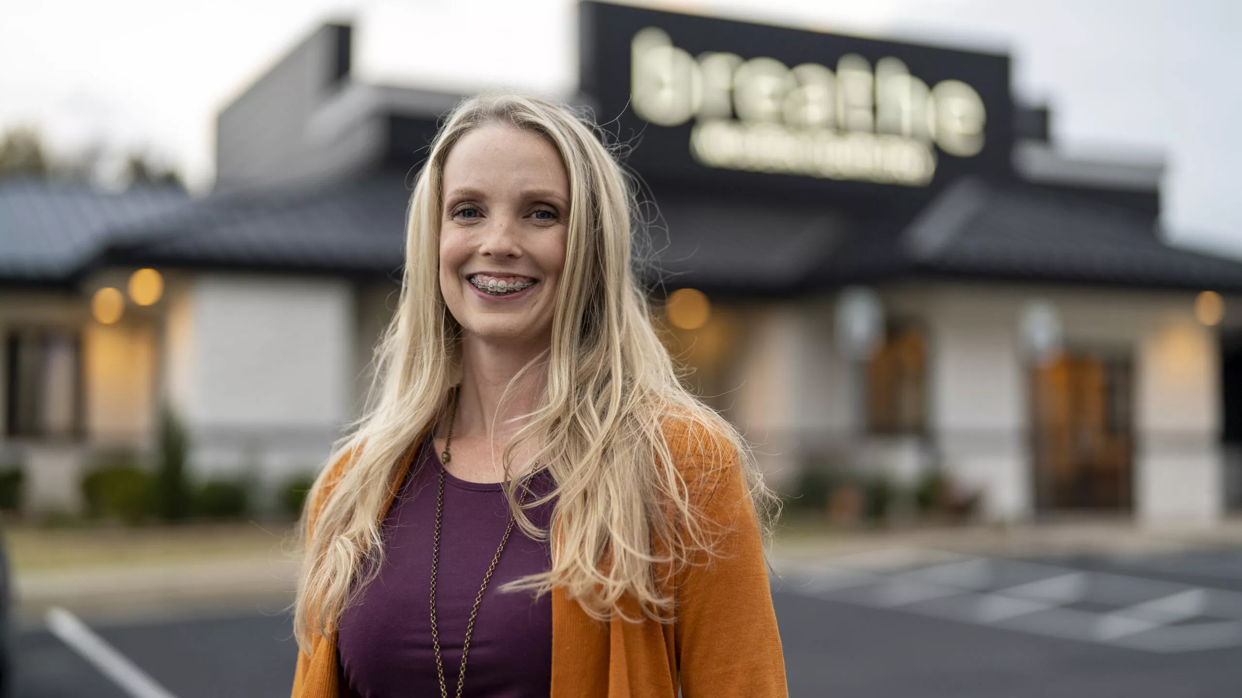 A woman with long blonde hair and braces is smiling and standing outdoors in front of a building with a sign that reads "breathe." She is wearing an orange jacket over a purple top, radiating positivity. The scene has the feel of a charlie patric photography moment.