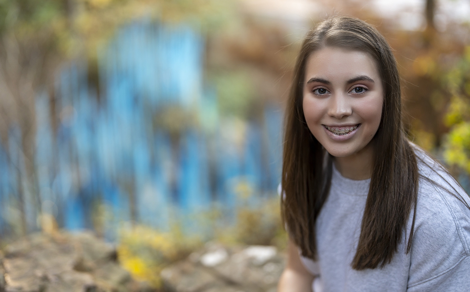 A young woman with long brown hair and braces is wearing a gray shirt and smiling in an outdoor setting with trees and greenery, perfect for high school senior portraits.