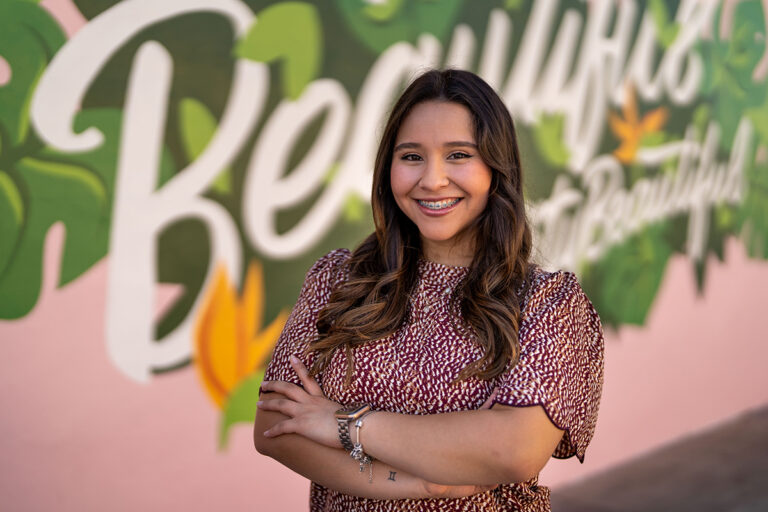 A woman with long hair and a patterned top stands smiling with arms crossed in front of a colorful mural with floral designs and text, capturing the vibrant spirit of high school senior portraits.