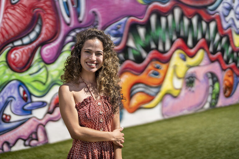 A woman with curly hair and a patterned dress stands smiling in front of a colorful, abstract graffiti wall, reminiscent of high school senior portraits.