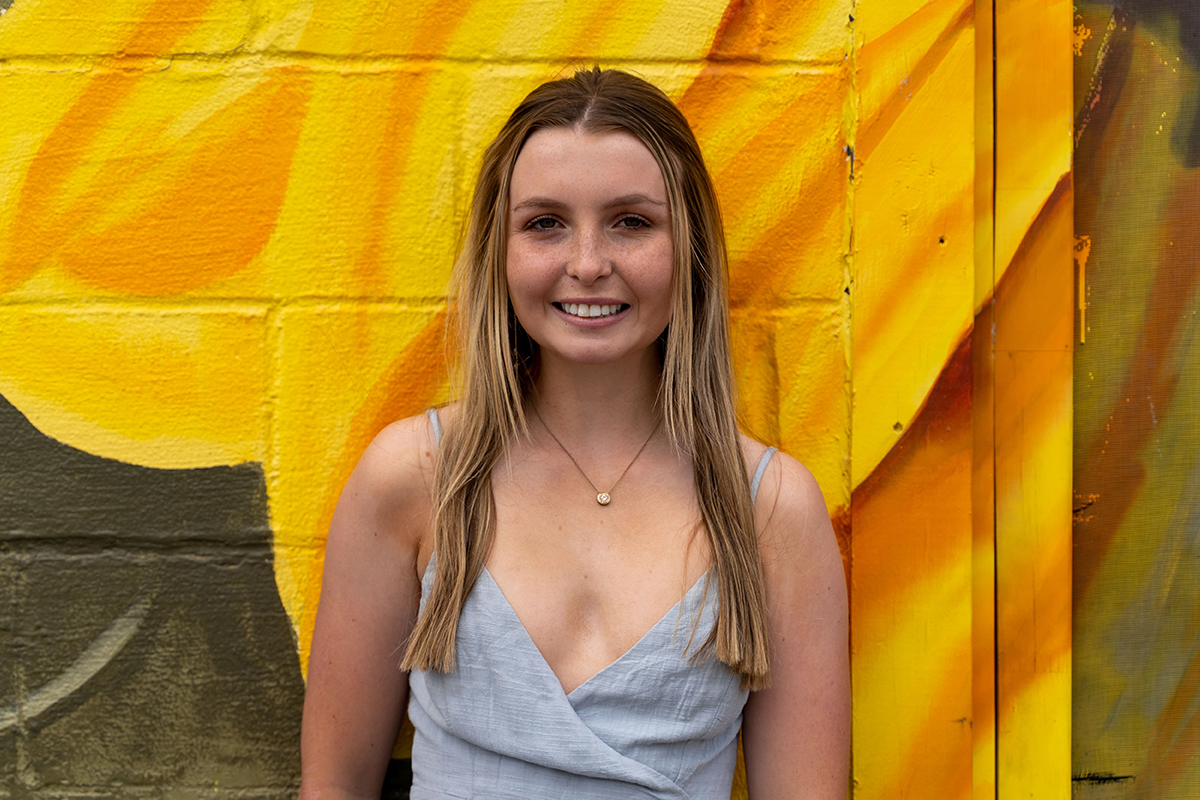 A woman with long blonde hair stands against a vibrant yellow and orange painted wall, smiling. She is wearing a light blue sleeveless top and a necklace, reminiscent of high school senior portraits.