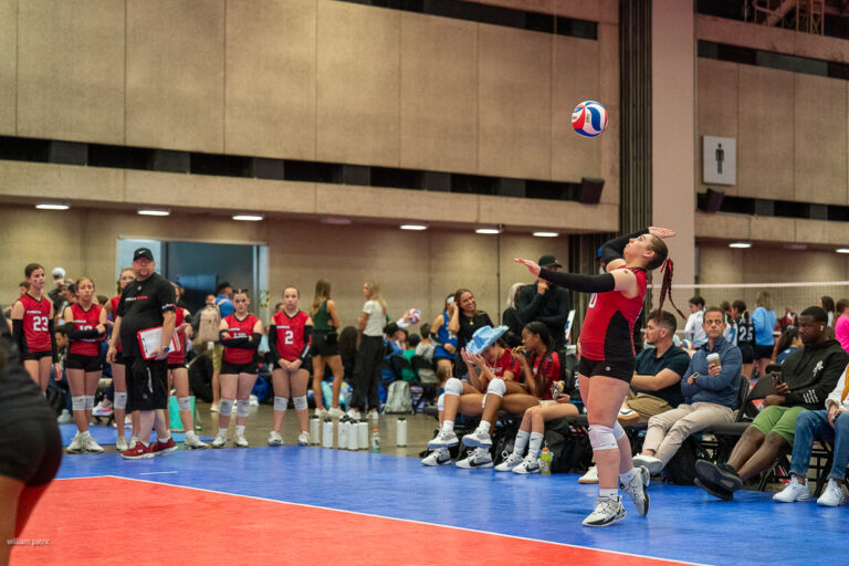 A volleyball player in a black and red uniform serves the ball during an indoor game. Teammates and spectators sit near the court's edge, observing the match.