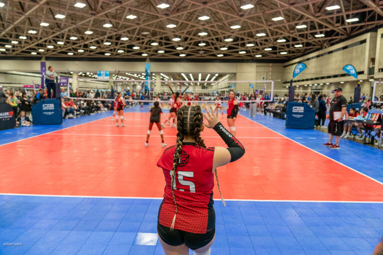 A volleyball player in a red jersey with the number 15 prepares to serve during an indoor volleyball match, set in a large brightly-lit gymnasium with spectators and other players in the background.