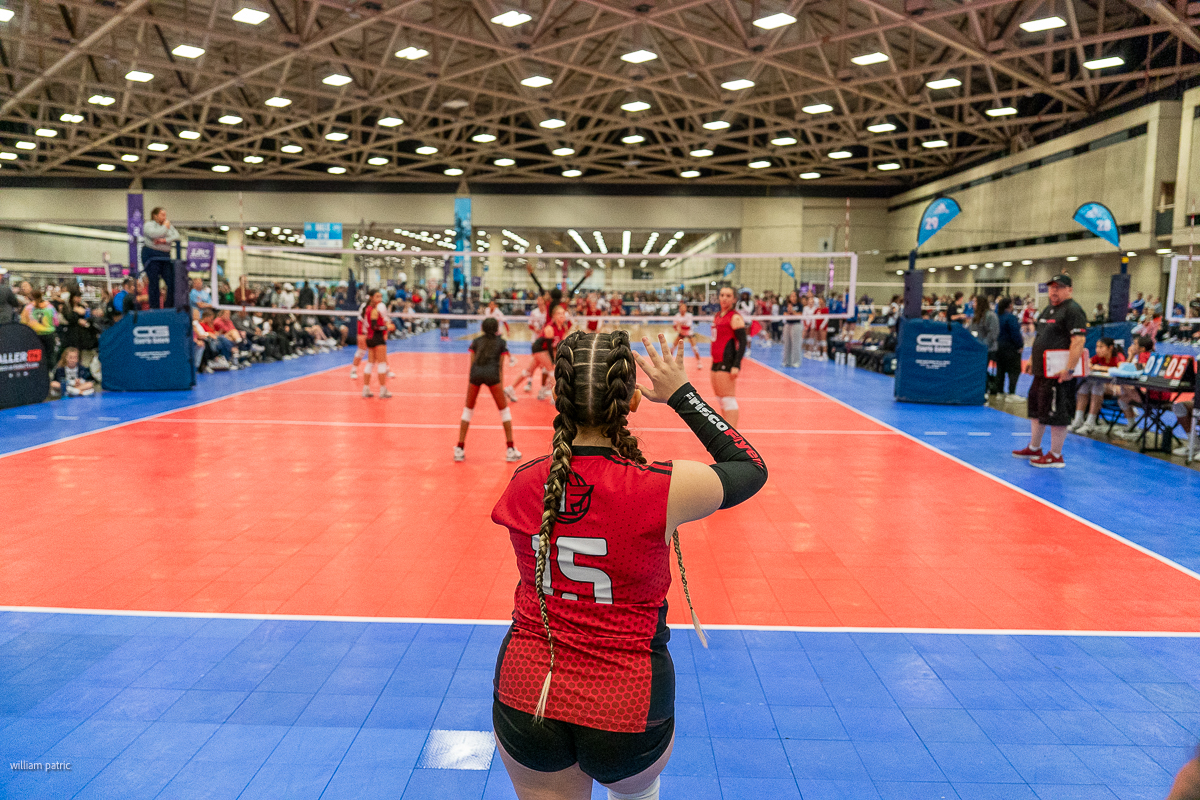 A volleyball player in a red jersey with the number 15 prepares to serve during an indoor volleyball match, set in a large brightly-lit gymnasium with spectators and other players in the background.