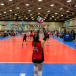 A volleyball player in a red jersey, number 15, prepares to serve during a match inside a large indoor sports facility.