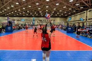 A volleyball player in a red jersey, number 15, prepares to serve during a match inside a large indoor sports facility.