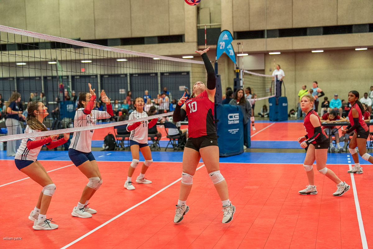A group of young athletes playing volleyball on an indoor court. One player in red (#10) is about to hit the ball while others prepare for the play. Spectators and officials are seen in the background.