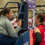 A volleyball referee in uniform speaks to a player in a red jersey number 10 during a game, with net and court in the background.