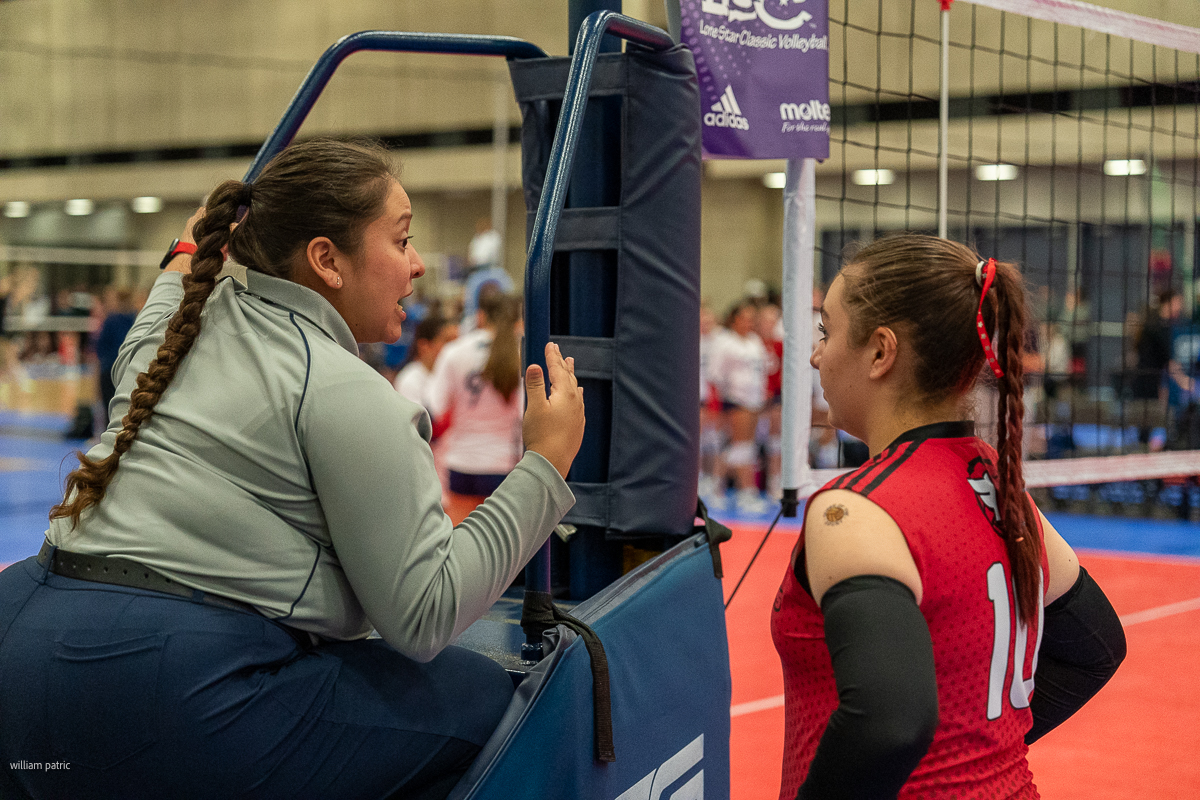 A volleyball referee in uniform speaks to a player in a red jersey number 10 during a game, with net and court in the background.