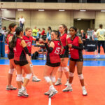 A group of six volleyball players in red jerseys stand together on an indoor court, engaging in discussion or strategy before a match. Spectators are visible in the background.