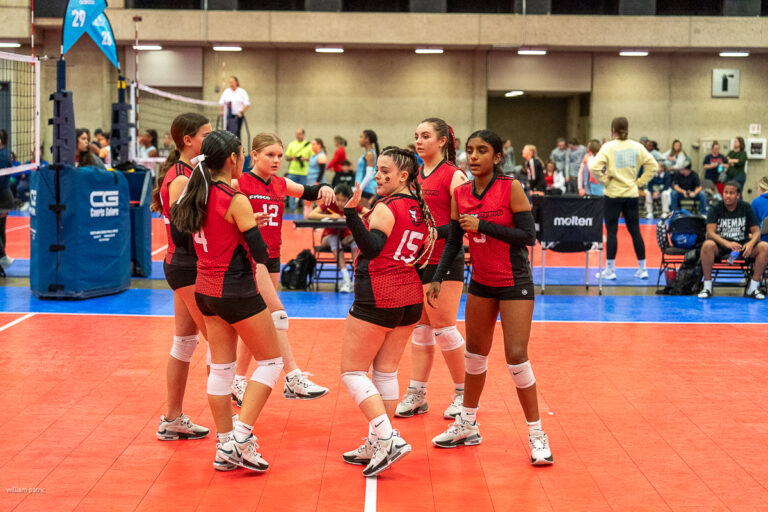 A group of six volleyball players in red jerseys stand together on an indoor court, engaging in discussion or strategy before a match. Spectators are visible in the background.