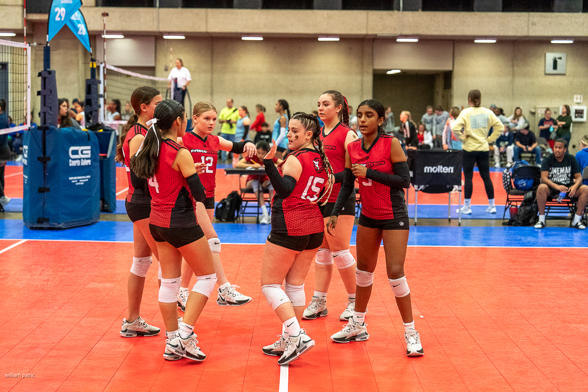 A group of six volleyball players in red jerseys stand together on an indoor court, engaging in discussion or strategy before a match. Spectators are visible in the background.