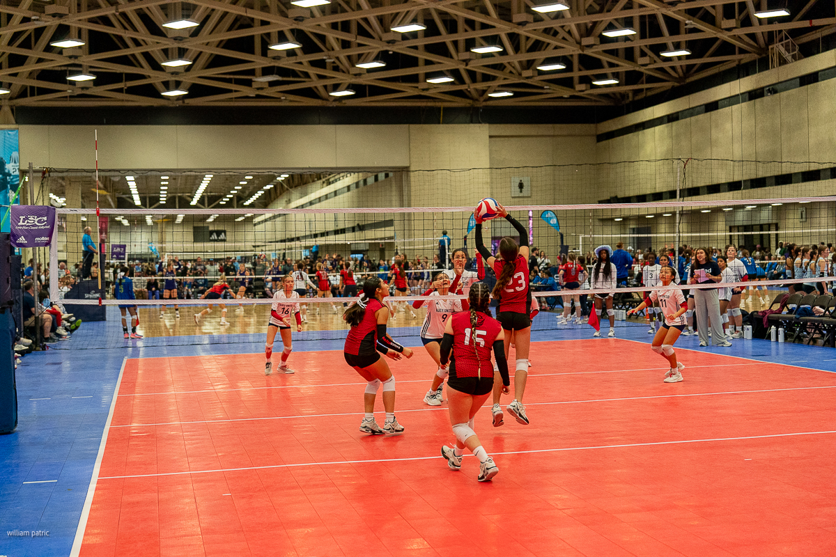 Indoor volleyball match with six players on each side, two players on the right team jumping for the ball. A crowded audience is watching from the stands in the background.