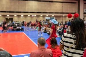 A woman wearing red headband pom-poms watches a volleyball game from the sidelines. A player in the background prepares to serve.
