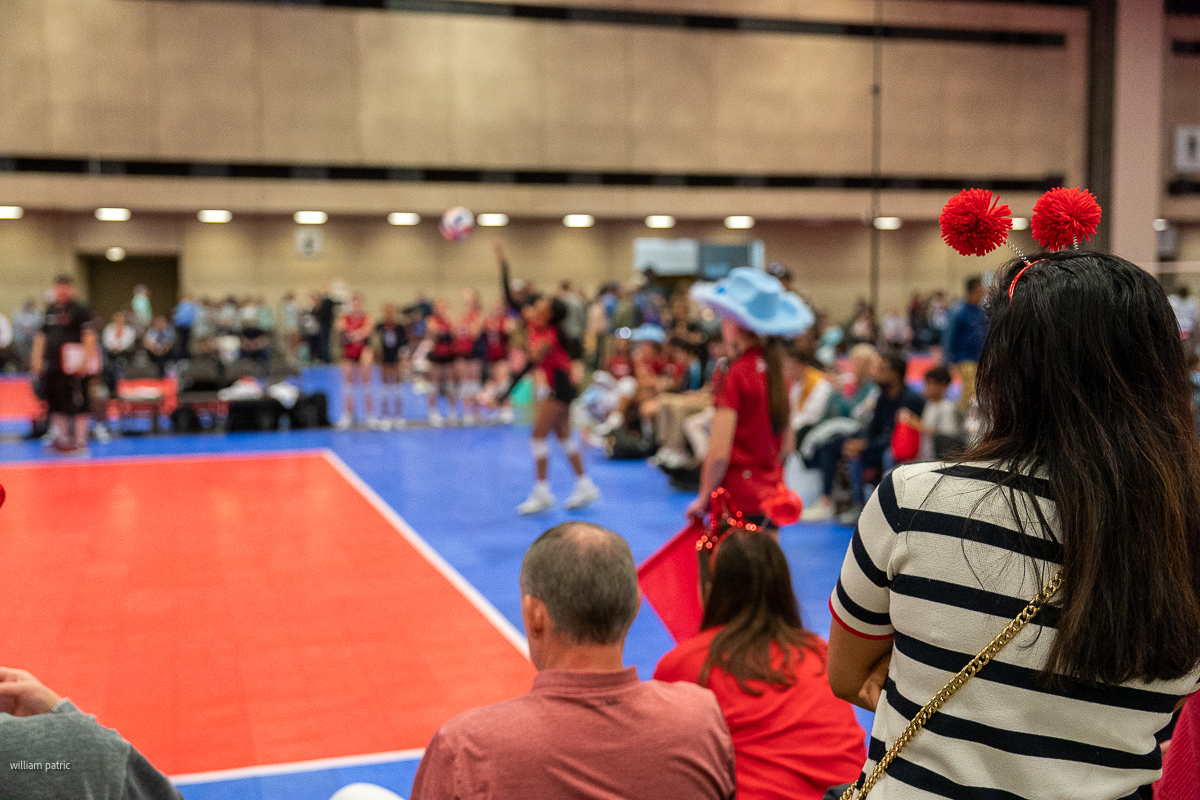 A woman wearing red headband pom-poms watches a volleyball game from the sidelines. A player in the background prepares to serve.
