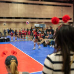 A volleyball player serves the ball in an indoor gymnasium, surrounded by teammates, spectators, and coaches.