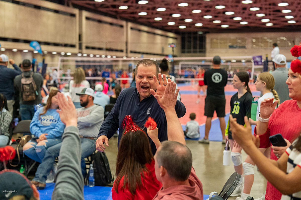 A coach enthusiastically high-fives supporters in the audience during an indoor volleyball game.