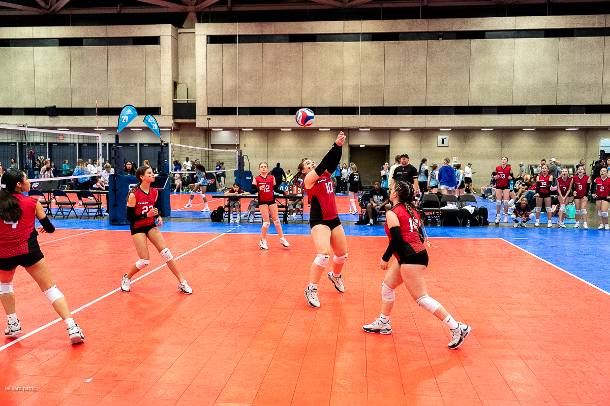 Indoor volleyball match in progress with team players in red jerseys actively competing. One player is jumping for the ball, while others prepare for the next move. Spectators and another team are visible.