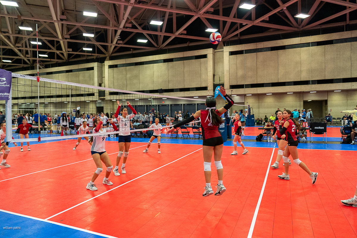 Indoor volleyball game in progress with players actively engaged on a red court under a high ceiling with bright lighting. Audience members and coaches stand in the background.