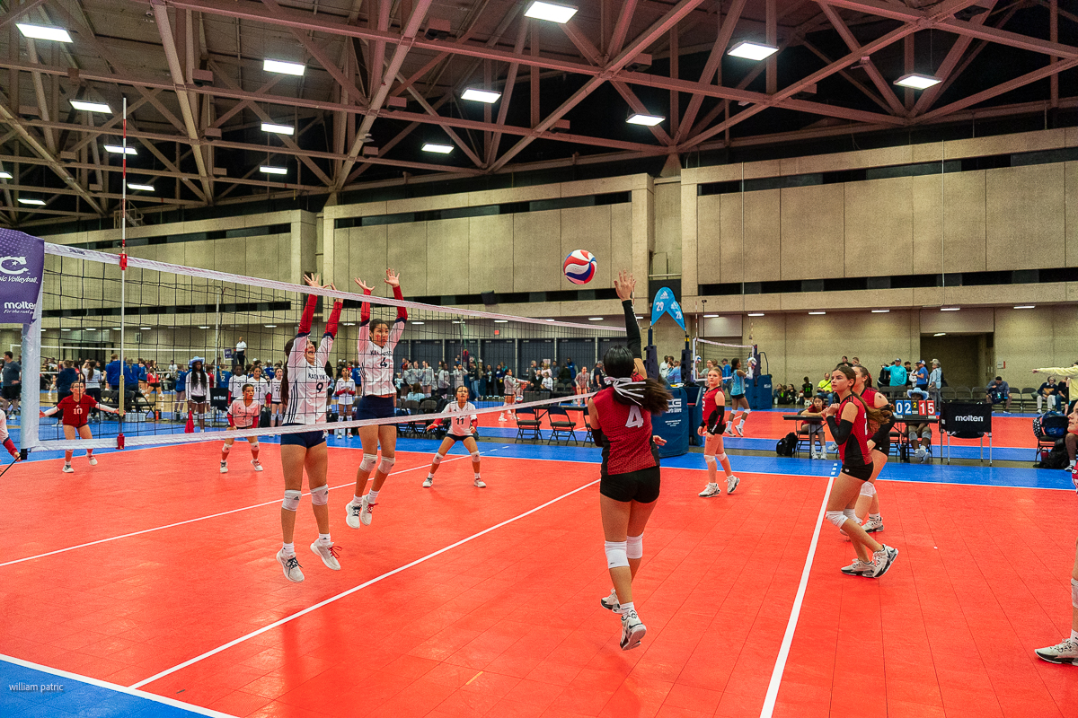 Indoor volleyball game in progress; players in white jump to block a spike from a player in red. The court is red, and spectators watch from the sides.