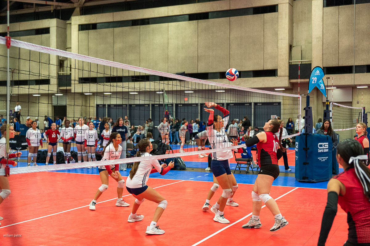 A women's volleyball match in an indoor court. One player in white and red is about to hit the ball over the net while teammates and opponents stand ready. Spectators and other players are in the background.