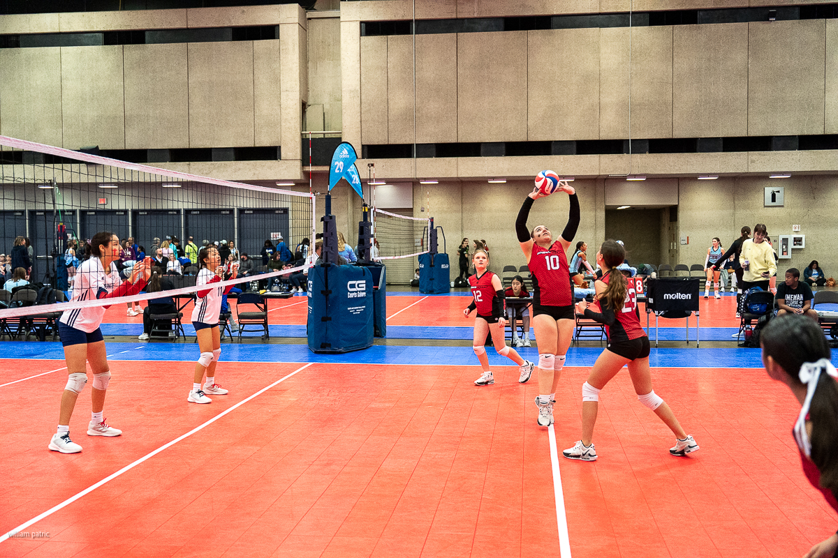 A volleyball game in progress indoors with six female players on each side. One player in red is setting the ball while her teammates and opponents are ready. Spectators and staff are visible in the background.