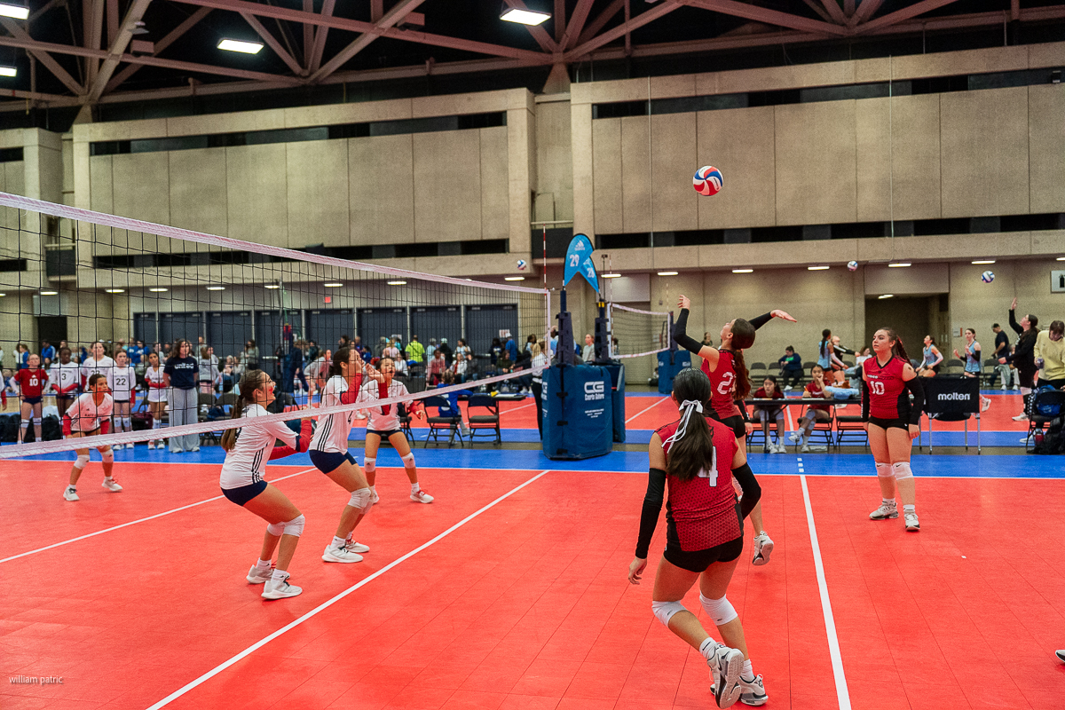 Indoor volleyball match with players in white and red uniforms. A player in red appears to spike the ball over the net, while others look on or prepare to respond. Spectators in background.
