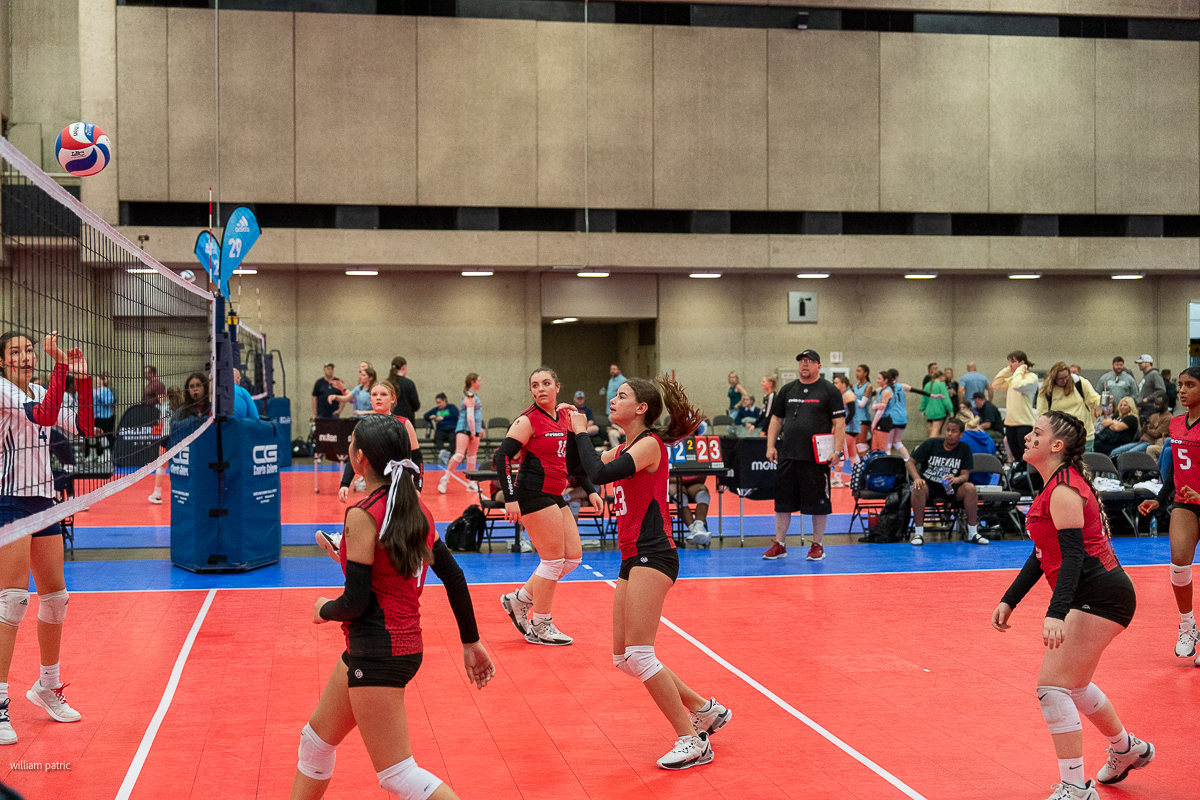 A women's volleyball team in red uniforms prepares to return the ball during an indoor match. The gymnasium has spectators and other players in the background.