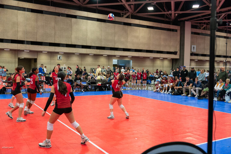 A women's volleyball match in progress on an indoor court, with players in red jerseys actively engaged and a crowd watching from the sidelines.
