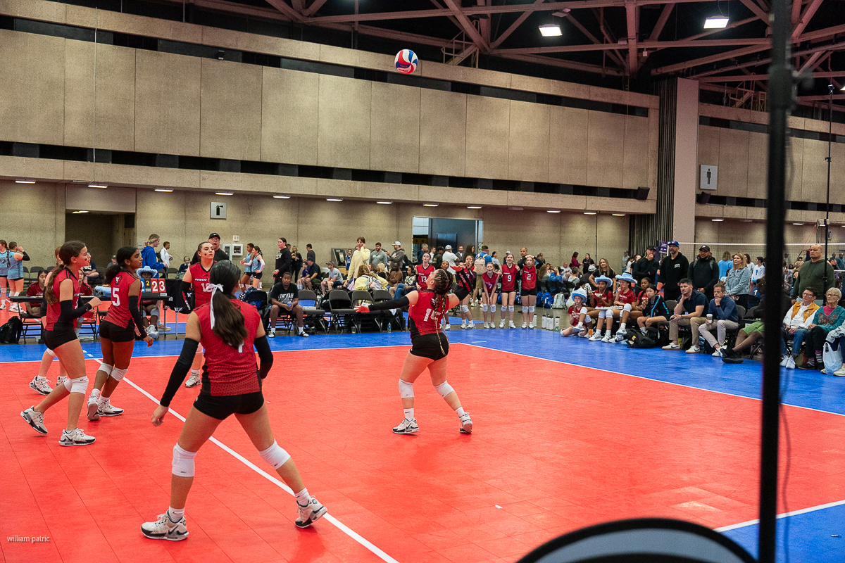 A women's volleyball match in progress on an indoor court, with players in red jerseys actively engaged and a crowd watching from the sidelines.