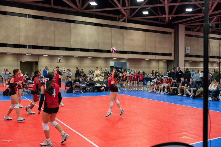 A women's volleyball match taking place in an indoor court with players in red and black uniforms in action, and spectators seated around the court watching the game.