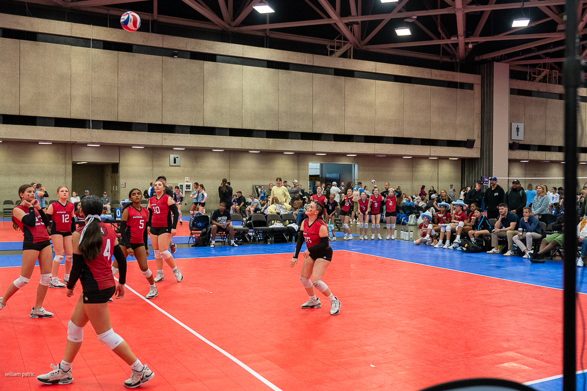 A women's volleyball team in red uniforms competes indoors, with spectators watching from the sidelines. A player prepares to set the ball while teammates are positioned around her.