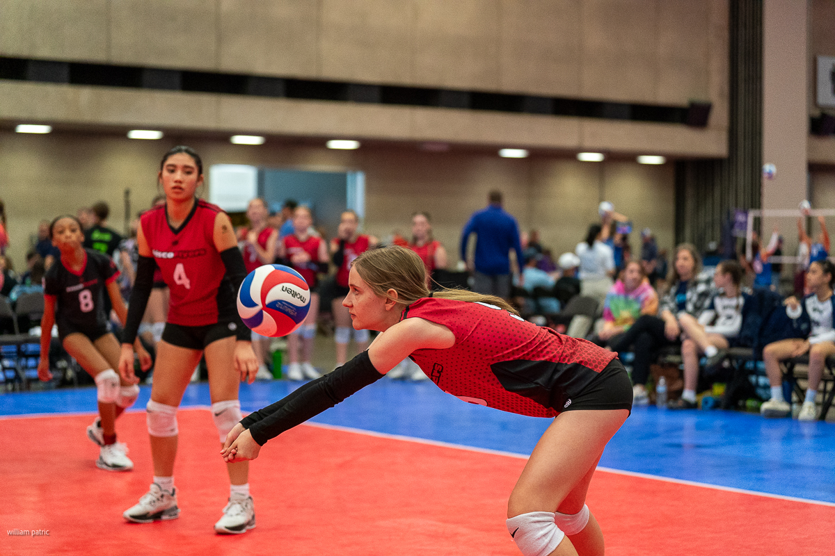 A volleyball player in a red jersey leans forward to bump the ball as a teammate and others watch in an indoor court setting.