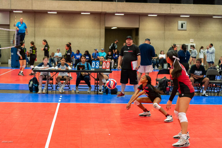 Two volleyball players in red jerseys prepare to receive the ball. A scoreboard shows 03-00. Spectators and officials are seated in the background.