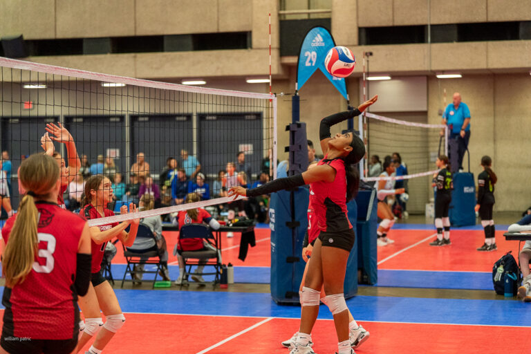 A volleyball player in a red jersey jumps to spike the ball over the net during a game. Other players and officials are visible in the background on an indoor court.