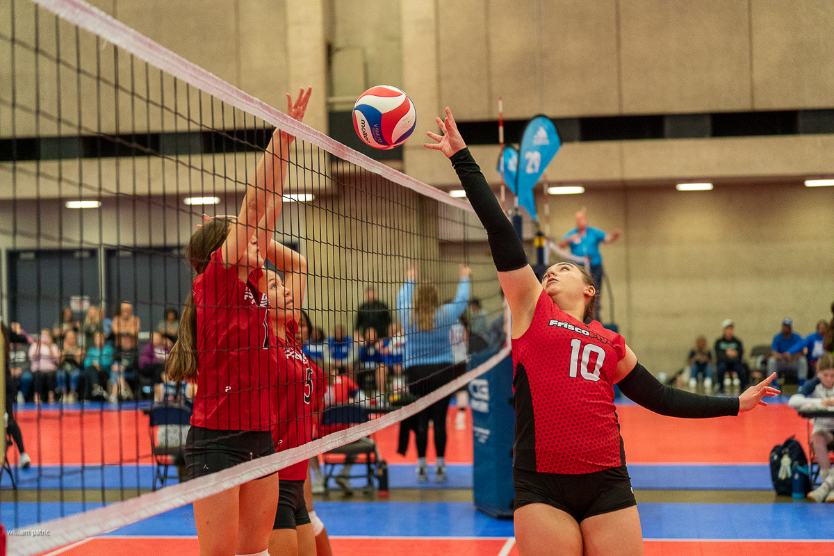 Indoor volleyball match with one player in a red jersey (number 10) hitting the ball over the net while two opposing players in red jerseys attempt to block.