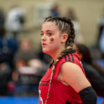 A female athlete with braided hair, wearing a red and black uniform, stands on an indoor sports court. She has stickers on her face and is looking to the side.