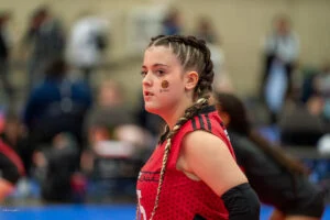 A female athlete with braided hair, wearing a red and black uniform, stands on an indoor sports court. She has stickers on her face and is looking to the side.