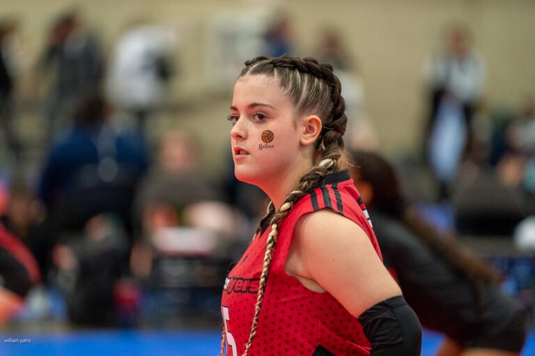 A female athlete with braided hair, wearing a red and black uniform, stands on an indoor sports court. She has stickers on her face and is looking to the side.