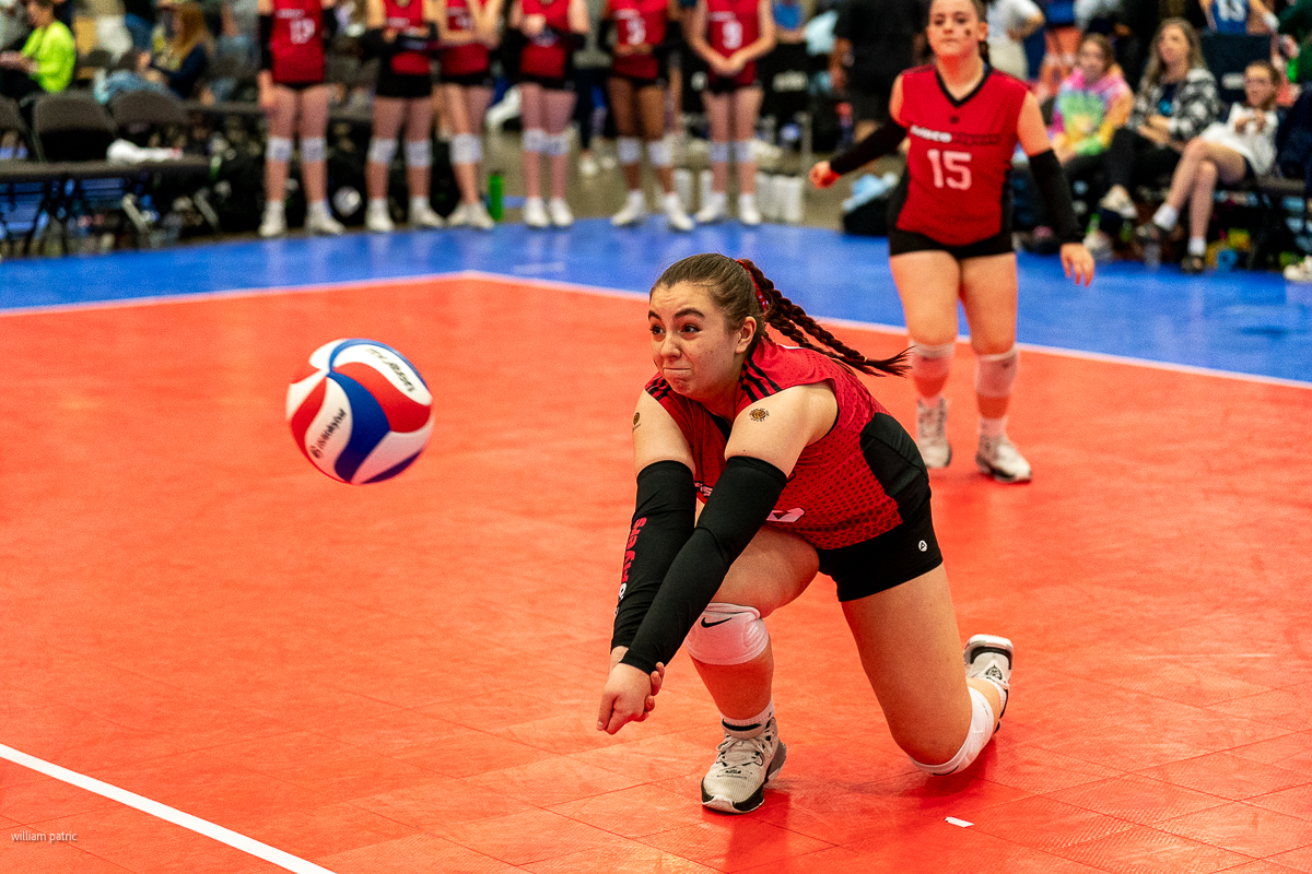 A volleyball player in a red uniform dives with outstretched arms to bump a ball during a game; another player is visible in the background.