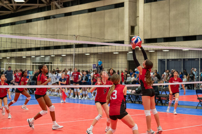 Indoor volleyball game with a player in a black and red uniform attempting to block the ball at the net, while teammates and opponents look on. Audience seated in the background.